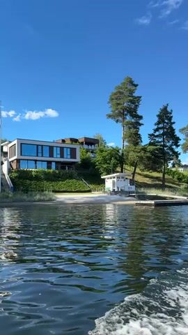 The video shows a body of water in the foreground with ripples and a wake from a boat. In the background, there is a shoreline with a sandy beach and some tall grass. A modern, multi-level house with large windows sits on a hillside behind the beach. To the right of the house, there is a small white building with a light-colored roof. Tall pine trees are visible on the hillside, and the sky is clear and blue. The perspective suggests the video is filmed from a moving boat approaching the shore.