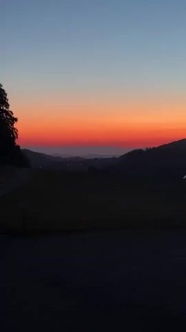 The video captures a serene sunset over a hilly landscape. The sky transitions from a pale blue at the top to a vibrant orange and red band near the horizon, indicating the sun is setting. Silhouetted against the colorful sky are the undulating shapes of distant hills, fading into a hazy purple in the distance. In the foreground, darker hills and trees are visible in silhouette, with a hint of a road or path on the left. The overall mood is peaceful and tranquil, with the natural beauty of the sunset dominating the scene. No people or specific objects are clearly identifiable, emphasizing the vastness and natural beauty of the environment.