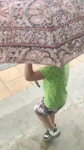 The media shows a close-up, eye-level view of a person walking under a large, patterned umbrella. The umbrella's fabric is intricately designed with paisley motifs in shades of red, pink, and brown against a dark background. The person beneath the umbrella is wearing a bright green t-shirt with a subtle pattern of what appear to be leaves or dots, and plaid shorts in red, black, and white. They are also wearing black and white sneakers. The person's legs and feet are visible, and they appear to be walking on a wet surface, possibly a sidewalk or street, indicated by the reflections and damp appearance of the ground. The umbrella is held open, sheltering the person from above. The background shows a yellow, textured surface, possibly part of a tactile paving system, and a grey concrete or asphalt ground. The lighting suggests it might be daytime, but the overcast or rainy conditions are implied by the wet ground and the use of the umbrella. The scene appears to be outdoors, on a pedestrian walkway. The individual's posture and the way they are holding the umbrella suggest they are moving forward, likely navigating the weather. There is no visible text in the media.