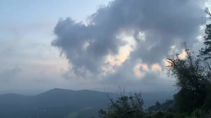 The video shows a landscape with mountains and clouds. The sky is filled with large, grey clouds, with some patches of lighter color, possibly sunlight, peeking through. In the foreground, there are trees and branches, some of which are bare. The mountains in the background are covered in a hazy, dark green color, suggesting distance. The overall atmosphere is muted and somewhat overcast, perhaps indicating dawn or dusk. There are no people or any discernible actions or interactions visible.