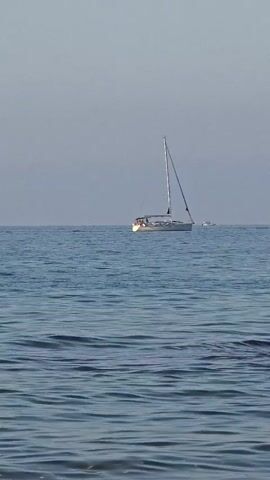 A sailboat is on the ocean. The sky is clear and blue. The water is calm with small waves. The sailboat is white with a tall mast. There are people on the deck of the sailboat. The scene is peaceful and serene.