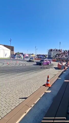 Two cars are drifting on a paved area. The car on the left is blue, and the car on the right is black and yellow. Both cars are kicking up smoke from their tires. Spectators are watching from a tiered seating area on the right. In the background, there is a large white tent, a building with an "AQVA" logo, and trees. The sky is clear and blue. The ground is paved with interlocking bricks, and orange traffic cones are placed around the drifting area.