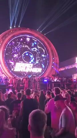 A large circular LED screen displays the name "BRAD BRUNNER" in large, stylized text, with abstract purple and blue visuals behind it. The screen is illuminated by bright lights and beams of light emanate from above, directed towards the sky. Below the screen, a stage is set up with performers and DJ equipment. A crowd of people are gathered in front of the stage, facing the screen and performers. The scene is lit by a combination of stage lights and the screen's illumination, creating a concert or festival atmosphere. The video captures a moment during a performance.
