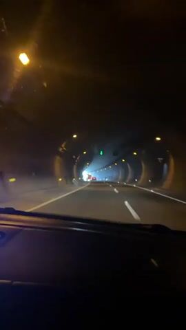 The video shows a first-person view from inside a vehicle driving through a tunnel. The tunnel walls are dark and lined with lights. There are other vehicles visible ahead, and the tunnel opens into daylight at the end. The camera is pointed forward, showing the road and the vehicle's dashboard in the foreground. The overall atmosphere is dark with a bright light at the tunnel's exit.