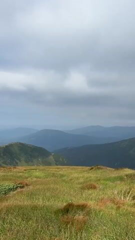The video opens with a wide shot of rolling, grass-covered mountains under a cloudy sky. As the video progresses, the view shifts to a mountain path with a lone hiker walking away from the camera. The hiker is carrying a large backpack and using trekking poles. The camera then pans across a vast, grassy meadow, revealing more undulating mountain terrain in the distance. The sky gradually clears, showing patches of blue and scattered white clouds. The final shot is a close-up of the grassy meadow, with the sky visible above.