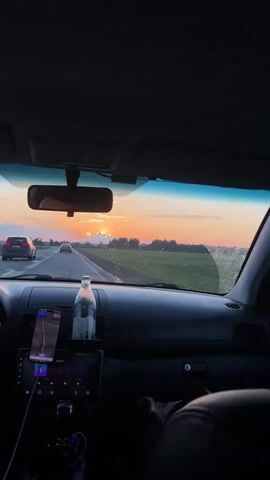 The video shows the interior of a car driving down a road during sunset. The camera is positioned behind the steering wheel, looking out through the windshield. The dashboard is visible, with a phone mounted on the left side displaying a navigation app. A bottle of water is placed on a shelf above the infotainment screen. The road ahead is lined with green fields. In the distance, the sun is setting, casting a warm orange glow across the sky, with some clouds visible. Several cars are also driving on the road ahead. The windshield has some water droplets on it.
