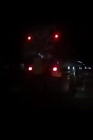 The video captures a large vehicle, likely a truck or bus, at night in Simpang, Indonesia. The scene is dark, with the vehicle's lights providing the primary illumination. Initially, the rear of the vehicle is visible, showing red taillights and several white/blue marker lights along its side. As the recording continues, the perspective shifts to a side view of the vehicle as it passes. Multiple white/blue lights are prominent along the side of the vehicle, casting reflections in the darkness. An engine sound is audible throughout the video, consistent with a moving, large vehicle.