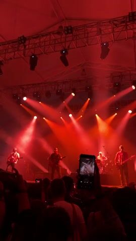 A band performs on a stage lit by red lights, with a dense crowd watching and recording on their phones. The band consists of four members playing guitars and drums. The stage is covered with a metal truss structure supporting numerous spotlights. The audience is mostly silhouetted against the bright stage lights, many holding up their phones to capture the performance. The overall atmosphere is energetic and immersive due to the vibrant lighting and the music.