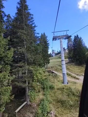 The video shows a view from a ski lift traveling upwards through a forest on a sunny day. The ski lift is supported by tall grey poles, and several ski lift cabins are visible moving along a cable. Dense green pine trees line the left side of the frame, with a dirt path visible winding through the grass and trees on the right. The sky is a clear blue with a few scattered white clouds. The overall impression is of a scenic mountain landscape during the summer or early fall.