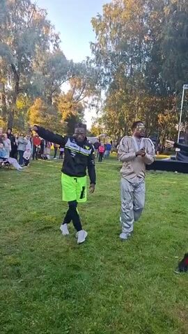 Two men are dancing on a grassy field in front of a stage. The man on the left is wearing a black jacket with yellow accents, neon green shorts, black leggings, and white sneakers. He has dark skin and is wearing glasses. The man on the right is wearing a light-colored, shiny jumpsuit and white sneakers. He has dark skin and is wearing glasses. Other people are visible in the background, some standing and watching, others on the stage. Trees and a building are also visible in the background. The scene appears to be an outdoor event or festival.