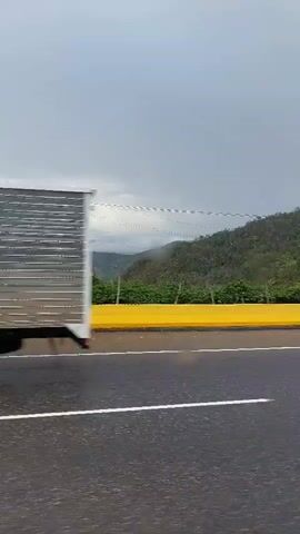 The video captures a view from a moving vehicle on a road, likely a highway, under an overcast sky. In the foreground, a dark asphalt road with a single white line is visible, appearing wet as if from recent rain. A bright yellow concrete barrier separates the road from the natural landscape. Beyond the barrier, lush green hills and mountains stretch into the distance, partially obscured by low clouds or mist, creating a layered effect. The vegetation on the hills is dense. Utility poles and power lines can be seen strung across the landscape. At the 0:02 mark, a small, light-colored car briefly passes by on the road. The overall scene suggests a journey through a mountainous, possibly rural or semi-rural, area during a cloudy day in a location like El Vapor, Venezuela.