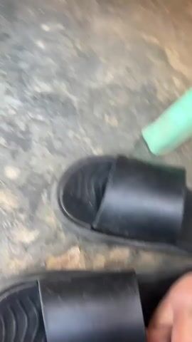 The video captures multiple individuals' feet and legs, primarily focusing on their footwear. Several people are wearing black slide sandals. One person's bare feet are shown before they put on a pair of black slide sandals. Another person is seen wearing green pants and black slides, while a third individual wears dark pants and black slides, with a watch visible on their left wrist. The setting appears to be outdoors on a textured concrete or dirt surface. This scene takes place in Kasuwan Talba, Nigeria.
