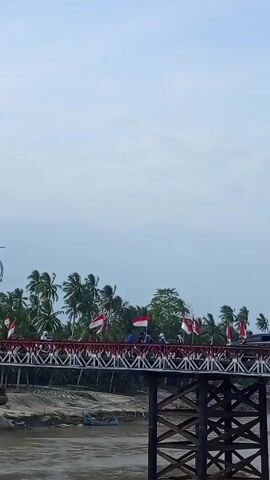 The video shows a medium shot of a red and white bridge decorated with Indonesian flags. People are visible on the bridge, and a car is driving across it. The bridge is supported by wooden structures, and a river flows underneath. Palm trees and other vegetation are visible on the riverbank. The sky is overcast. Loud sound is also audible.