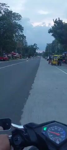 The video shows a POV shot from a motorcycle traveling down a two-lane road. The road is paved and has a distinct line down the middle, separating what appears to be two different paving materials or conditions. On the left side of the road are trees and some buildings, with a few motorcycles visible. On the right side of the road, there are trees, utility poles, and several parked vehicles, including yellow tricycles and a white pickup truck. The sky is cloudy and overcast, suggesting it is either dawn or dusk. The overall impression is that of a rural or semi-urban road in a tropical or subtropical region.