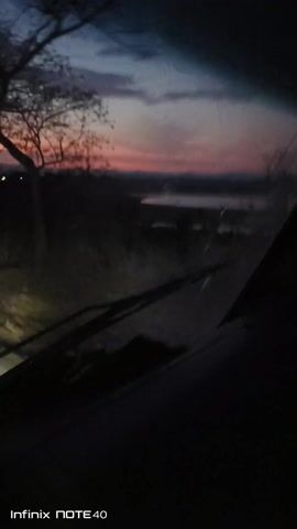 The video shows a view from inside a vehicle, looking out through the windshield as it travels along a road at dusk or dawn. The sky is visible with a gradient of colors, ranging from blue to orange and pink hues, suggesting a sunrise or sunset.  Silhouettes of trees are seen on the left side of the frame against the colorful sky. The road ahead is illuminated by the vehicle's headlights, and other distant lights are visible in the background. The windshield appears to have some cracks or imperfections. The vehicle is in motion, creating a sense of forward movement.