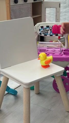 A yellow plastic duck toy with an orange beak and feet, standing on a white wooden children's chair with light wooden legs, is the central focus of a video. In the background, to the left, is a white wall with a black polka dot pattern. To the right, a pink and white plastic toy ice cream cart with the words "ice cream cart" on its side is visible. The floor is light grey, and a light-colored rug is partially visible beneath the chair. The duck remains stationary throughout the video, and there are no people visible.