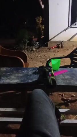 The video opens with a close-up of someone's legs in grey sweatpants, resting on a wooden railing. The camera then pans up to reveal a dark outdoor scene. In the foreground, a weathered blue table is visible, with a glass of dark liquid and a pair of sandals resting on it. To the right of the table is a green plastic bucket and a pink object. In the background, there's an orange plastic chair, and beyond that, an area with dirt and some foliage. A white building with a dark window frame is on the far right. A vehicle with illuminated headlights drives by in the distance, briefly lighting up the scene. The overall impression is a low-light, casual outdoor setting at night.