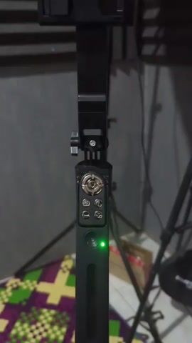 The video shows a close-up of a black selfie stick with a green light indicator on. The selfie stick has several buttons on it, including a circular directional pad, a camera icon, and other smaller buttons. The selfie stick is mounted on a tripod, and a colorful mat is visible on the floor in the background. The lighting is dim, and the focus is on the selfie stick.