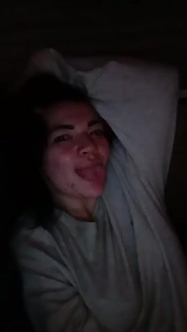 The video shows a person with shoulder-length dark hair wearing a long-sleeved grey shirt, lying down. The person briefly sticks their tongue out and then smiles. The lighting is dim, and the background is dark. The person appears to be indoors.