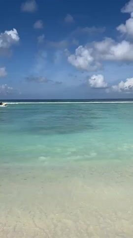 A jet ski with two riders is speeding across the turquoise water. The jet ski is in the distance, and the riders are small figures on the vehicle. The water is clear and blue, with some white foam from the jet ski's wake. The sky is blue with some fluffy white clouds. The shoreline is visible in the foreground, with light-colored sand and shallow water. The scene is a tropical beach on a sunny day.