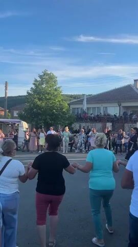 The video shows a group of people dancing in a circle, holding hands, in a street with houses and trees in the background. The sky is blue with some clouds, and the lighting suggests it is either early morning or late afternoon. Some people are watching from the balconies of the houses. One man in a blue polo shirt is clapping, possibly in time with the music. The overall atmosphere appears to be festive and communal.