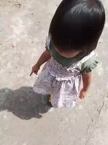 A young child wearing a green top, a white floral-patterned dress, patterned leggings, and yellow sandals stands on a cracked concrete path outdoors. The child's hair is dark and cut into a bob. The child's shadow is visible on the ground to their left. The child looks at the camera briefly, then looks to their right, and then to their left. The background shows a slightly elevated area on the left and a wider path extending into the distance on the right, with some foliage visible in the distance. The lighting suggests it is daytime.