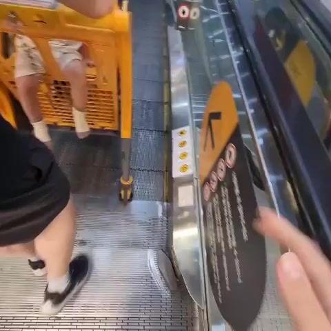 The video begins with a close-up of a child's legs sitting in a yellow shopping cart as it ascends an escalator. The camera then pans to the side, showing the escalator steps and the handrail. A person's arm is visible on the right side of the frame, holding a phone. As the video progresses, the escalator continues to move upwards, revealing more of the surrounding environment, which appears to be a shopping mall. Other shoppers and carts can be seen on the escalator. The child in the cart looks around and appears to be engaged by something off-camera. The video ends as the escalator continues its ascent.