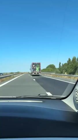 A first-person perspective video shows a semi-trailer truck on a highway under a clear blue sky. The truck's rear doors are decorated with an image of two characters. On the left is a woman dressed in a black, ornate outfit, holding a red apple and looking at the viewer. On the right is a green-skinned character with dark hair and a black hat, appearing to be looking forward. The truck has a license plate that reads "DR-56-ZJ." The highway has multiple lanes, and other vehicles are visible in the distance. The environment appears to be rural with trees and vegetation lining the sides of the road.