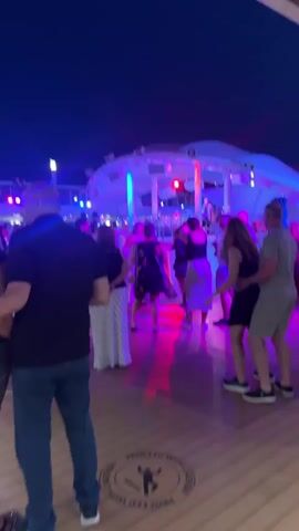 A group of people are dancing at night on a wooden deck, illuminated by colorful lights from a stage in the background. The stage has a DJ booth and speakers, and several people are visible on or near it. Some of the dancers are in the foreground, facing away from the camera. The mood appears festive and energetic.