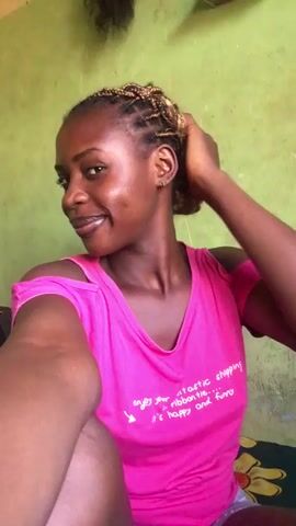A medium shot shows a young Black woman wearing a pink cut-out shoulder t-shirt. She has dark skin and light-colored braids pulled back from her face. She is smiling and looking at the camera. She is sitting indoors, and the wall behind her is painted a pale green color. She raises one hand to the camera. Her teeth are very white and her lips are shiny. The camera moves slightly and focuses on her. The woman strikes a thinking pose, with one hand under her chin. The video blurs a bit near the end.