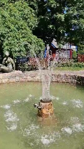 This media file captures a tranquil outdoor scene featuring a circular fountain with water jets. In the background, a lush green tree canopy provides shade, and a rustic building with red siding and blue trim, reminiscent of traditional European architecture, is visible. A stone wall surrounds the fountain, and a bronze sculpture of a reclining figure is positioned behind it to the left. To the right, a wooden bench rests on the grass. The sunlight suggests it is a bright, clear day, likely daytime. The overall atmosphere is peaceful and serene, evoking a sense of calm and natural beauty. The location is identified as Neringa, Lithuania. There is no visible text in the media file.