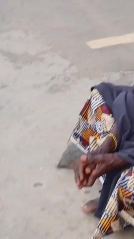 A person is sitting on the ground with their legs crossed and hands clasped. They are wearing a patterned skirt and a dark gray garment. The ground appears to be a concrete surface. The camera moves away from the person, focusing on the ground. There is faint background noise, possibly voices.