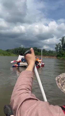 The video begins with a first-person perspective of someone holding a paddle on a body of water. In the distance, a raft carrying four people is visible. The raft appears to be made of inflatable tubes and wooden planks. Some of the people on the raft are wearing life vests. The environment consists of a river with tree-lined banks and a cloudy sky. The person holding the paddle seems to be rowing or steering. The raft in front is moving in the same direction. The scene suggests a recreational activity like rafting or kayaking.