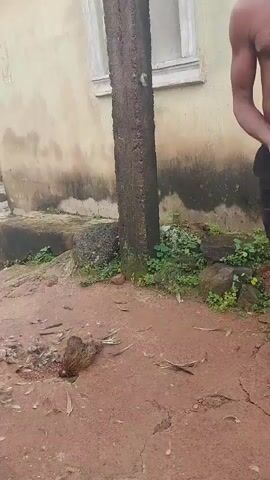 A video shows a bird lying dead on the ground near a wooden pole. A man stands to the right of the pole, holding something in his hand. Another person, partially visible, stands to the far right. The ground is dirt and gravel, with some green foliage. A building with a light-colored wall and a window is in the background. A second clip shows the same scene, with the man now more fully visible, shirtless and wearing dark pants. He appears to be holding small objects in his hand. In a third clip, a young person wearing a blue cap and a patterned jacket sits near a wall. A fourth clip shows a group of people in what appears to be an outdoor setting. One person, wearing a brightly colored shirt, stands and holds a small object. Another person, lying down, holds what looks like a cigarette. In the background, a fire is burning, with smoke rising. There are green leaves on the ground.