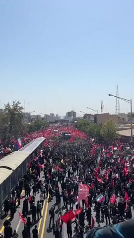 A large crowd of people are gathered in a street, many holding red flags and some holding Iranian flags. There are several vehicles, including a truck with a banner on its side, moving through the crowd. Buildings and trees are visible in the background under a clear blue sky. The scene appears to be a procession or a demonstration.