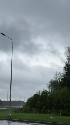 The scene shows an overcast sky with thick, grey clouds dominating the upper portion of the frame. Below the sky, a row of evenly spaced streetlights lines a highway. To the right, a dense line of green bushes and trees obscures part of the horizon. Rain appears to be falling, creating a sheen on the ground. A few cars are visible on the highway.