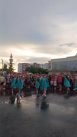 This media file captures an outdoor event in Kryvda Nivka, Russia. The scene features a group of young girls performing a dance routine on a wet, asphalt surface, likely a public square or street. They are dressed in matching turquoise dresses that flow as they move. A large crowd of onlookers, composed of adults and children, stands behind the performers, observing the event.

The setting includes several multi-story buildings in the background, indicative of an urban or semi-urban environment. A tall evergreen tree is visible to the left of the frame. The sky is overcast with a soft, diffused light, suggesting it might be late afternoon or early evening. The wet ground, with visible reflections, indicates recent rain or moisture. The overall atmosphere appears to be one of community gathering and entertainment, with the spectators appearing engaged and possibly enjoying the performance. There is no visible text in the media file.