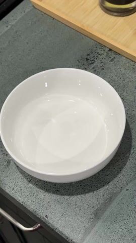 A hand pours the contents of an orange and white package of “Baker at 99 Simple Golden Syrup Flavour 100% Wholegrain Oats” into a white ceramic bowl. The bowl sits on a gray countertop. A wooden tray and a black object are visible in the background.