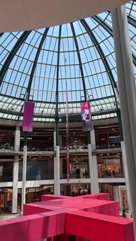 The video shows the interior of a large, multi-story shopping mall with a domed glass ceiling. The camera is positioned on an upper level, looking down towards the ground floor and up towards the ceiling. In the foreground, a large, bright pink, cross-shaped structure is visible. This structure appears to be an exhibit, with panels displaying text and images on its surfaces. Several people are visible walking around and interacting within the shopping mall, some on the ground floor and others on the upper levels. Stores and restaurants, including a McDonald's, are visible on the different levels. Large digital screens are suspended from the ceiling, displaying various images and advertisements. One screen shows a close-up of a person's face. Another screen displays a red rectangular graphic. The overall environment suggests a busy, modern commercial space.