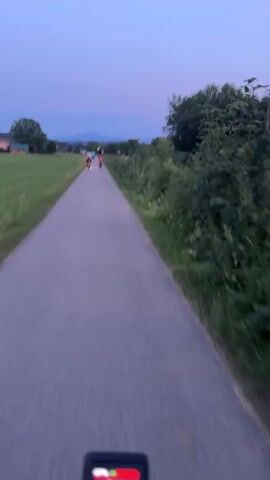 The video shows a first-person perspective of someone riding a bicycle on a paved path. The time display on the bike's handlebars indicates a speed of 10. The path is surrounded by green fields on the left and trees and bushes on the right. In the distance, there are a few buildings and mountains visible under a twilight sky. Further down the path, two people are riding bicycles, one of whom is pulling a small cart with another person inside. The camera is moving forward, capturing the scenery as the cyclist rides.