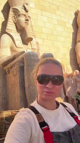 A woman wearing sunglasses, a white shirt, and a harness poses for a selfie in front of two large statues of seated pharaohs. The statues are made of a light tan material and are adorned with hieroglyphics. The background is a tan brick wall.  Behind the statues are additional smaller statues. In the distance are buildings and a blue sky.  There is a black iron fence in front of the statues.  The video pans from left to right to show the statues and then back to the woman.