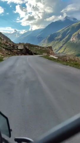 The video begins with a point-of-view shot from a moving vehicle, likely a motorcycle, as indicated by the handlebars visible in the foreground. The vehicle is traveling on a paved road that curves to the right, cutting through a mountainous landscape. Steep, rocky slopes covered with sparse vegetation rise on either side of the road. In the distance, majestic, green-covered mountains stretch towards a sky filled with dramatic, white clouds illuminated by sunlight. A single pole with what appears to be electrical wires is visible on the left side of the road. The road surface is a dark asphalt, showing some wear and tear. The overall impression is one of a scenic drive through a rugged, natural environment.