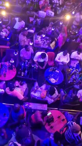This video captures a lively indoor/outdoor venue, likely a bar or club, from a high-angle perspective looking down on the patrons. The setting is illuminated by numerous string lights and ambient blue and purple lighting, suggesting it's nighttime.

The venue is bustling with a large crowd of people, mostly adults, seated at small round and square tables or standing and socializing. Many tables have bottles, glasses, and shisha pipes, indicating a lounge or bar atmosphere. People are engaged in conversations, using their phones, and generally enjoying the vibrant environment. Some individuals are seen moving to the rhythm of the upbeat music playing in the background.

The space appears to have multiple levels, with a balcony or upper floor visible in the background, also occupied by patrons. The overall mood is festive and social. One person wears a white shirt with "Aba n'anya" or similar text visible. The location context is Obosi, Nigeria.