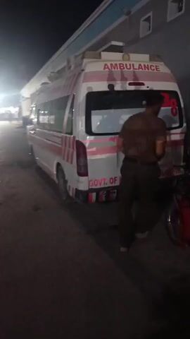A white ambulance with red stripes and "AMBULANCE" written on the back stands parked. The text "GOVT. OF KHYBER PAKHTUNKHWA" is also visible. The side of the ambulance reads "RESCUE 1122." A man is standing near the ambulance and a motorcycle is parked next to him. The scene appears to be at night.