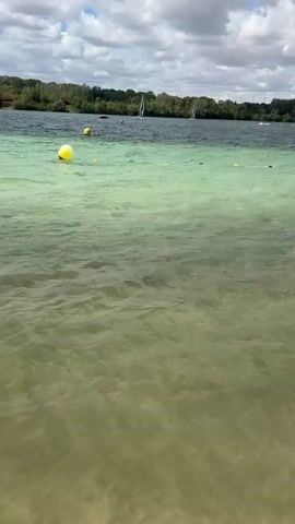 The video captures a body of water with a shoreline visible in the distance, lined with trees. The water exhibits a clear division in color, with a lighter, greenish hue in the foreground and a darker, blue-gray hue further out. Yellow buoys are scattered in the water, marking an area. Several sailboats are visible on the darker water, along with what appear to be individuals engaged in watersports. The sky is cloudy with patches of sunlight breaking through. The camera is positioned close to the water's surface, with gentle ripples present.