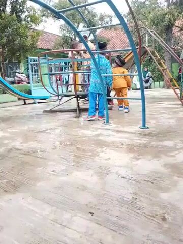 This video shows an outdoor playground scene in Kecamatan Gegesik, Indonesia, featuring several young children at play. In the foreground, a group of boys is actively engaged with a blue and red merry-go-round. One boy, dressed in a white and black plaid shirt and black pants, is seen running around the outside, pushing the merry-go-round, while another boy in a brown/orange outfit stands near it, then runs past it. Other children are also positioned on or around the merry-go-round as it spins.

The playground includes various colorful equipment, such as a yellow slide and a blue arched climbing structure, visible in the background. The ground is a concrete surface, which appears somewhat damp. In the background, behind the playground equipment, several adults are seated, observing the children. A building with a red-tiled roof and green elements is also visible in the distance, suggesting a school or public facility. The video is recorded during the daytime, under bright, possibly overcast, lighting conditions. The children appear energetic and to be enjoying their playtime.
