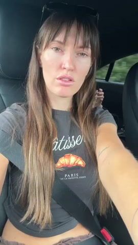 The video shows a woman with long brown hair and bangs, wearing a dark t-shirt with a graphic of a croissant and the word "Patisserie" and "Paris" on it. She is sitting in the driver's seat of a car, with a seatbelt across her chest. Sunglasses are perched on top of her head. The interior of the car is dark, and the background is blurred, suggesting motion. The woman is speaking directly to the camera, at times smiling and at other times opening her mouth wide as if singing or exclaiming. Her facial expressions change throughout the clip.