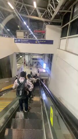A group of people are riding an escalator down to a lower level of what appears to be a train station or airport. The person recording is also on the escalator, behind the others. The camera is angled downward, showing the escalator steps, the backs of the people in front, and the space below. A sign above the escalator indicates directions to the "Airport" (left) and "Exit" (down). The lower level has a bright, open space with people walking around. The overall lighting is bright, with a slightly industrial feel due to the metal structure above.