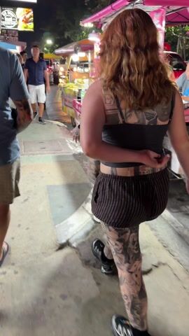 The video captures a dynamic night scene on what appears to be a busy street or market area, brightly lit by artificial lights. The camera primarily focuses on a person from behind, who has long, wavy reddish-brown hair. This individual is extensively tattooed on their back, arms, and legs.

The person is wearing a dark grey tank top and dark brown shorts with thin, light-colored vertical stripes. On their upper back, two prominent black and white portrait tattoos are visible, partially obscured by the tank top straps. One tattoo distinctly depicts a man's face, resembling Tupac Shakur, with another figure alongside it. Additional tattoo designs are visible just above the waistband of their shorts, underneath the tank top. Both arms and legs are heavily adorned with various black and grey tattoo artwork.

They are walking on a paved surface, likely a sidewalk, amidst a crowd of other pedestrians. To their immediate right, another person, also with visible arm and leg tattoos, wearing a light-colored top and shorts, walks slightly ahead. In the broader background, several brightly lit street stalls or food vendors contribute to the vibrant atmosphere. A man in a blue shirt and light shorts is visible walking to the left. The scene conveys the lively ambiance of an evening street, possibly a market or a popular tourist spot. No specific text or location identifiers are clearly discernible.
