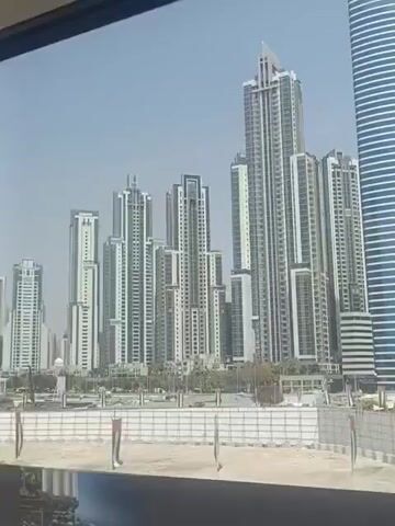 The video depicts a view of a city skyline from a moving vehicle, likely a train. The skyline consists of numerous high-rise buildings, predominantly white or light-colored with some dark accents. They are densely packed together, suggesting a major urban center. A barrier, likely construction fencing, is visible in the foreground along with sandy ground. The sky is clear and blue, indicating a sunny day. The camera angle is slightly elevated, looking out and across at the buildings. There are no visible people in the video. The video ends abruptly, with a shift in focus to an interior view.